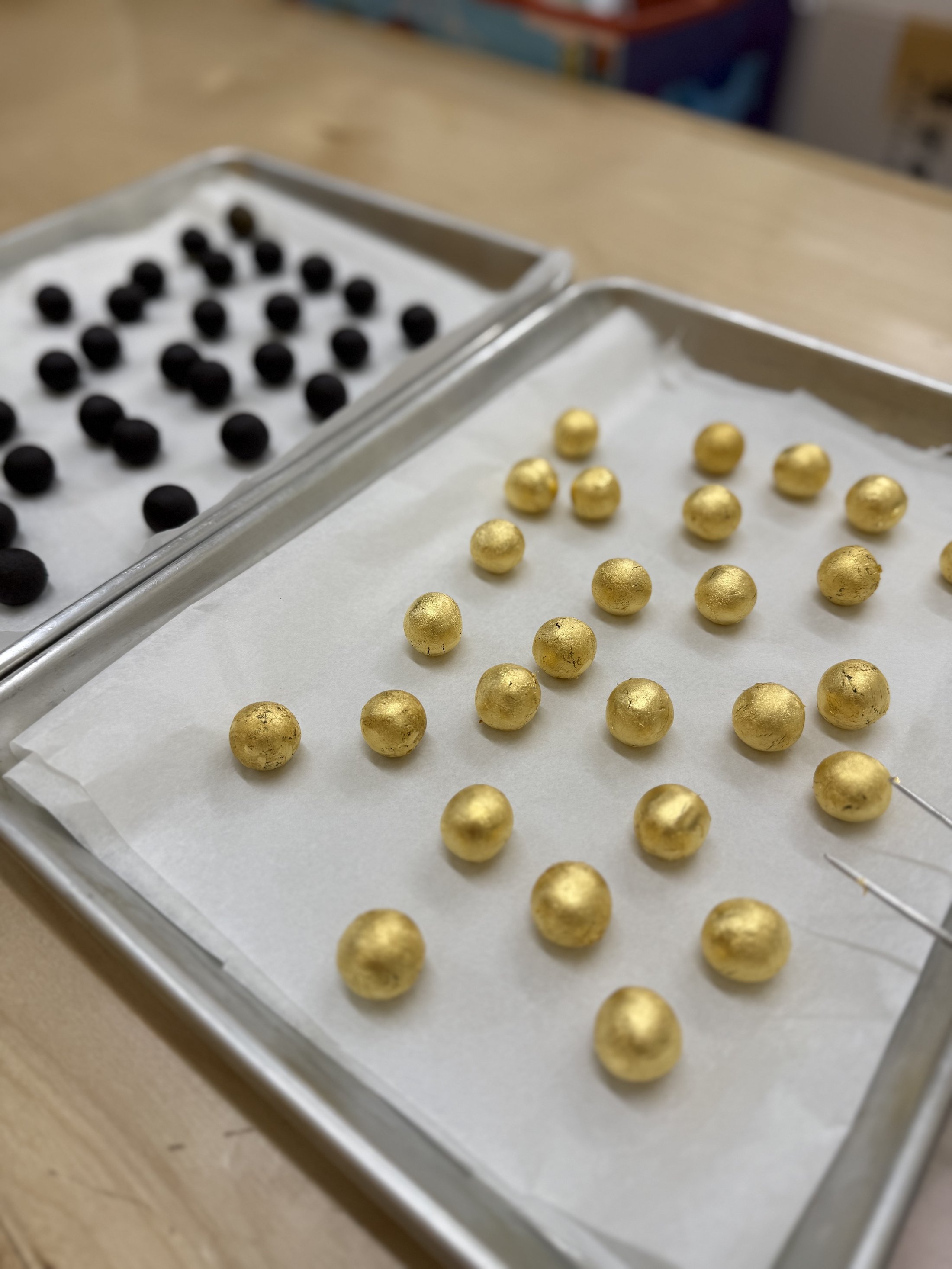 Trays of black charcoal-coated and gold-leaf herbal pills laid out on parchment, mid-process in the Harbin back kitchen