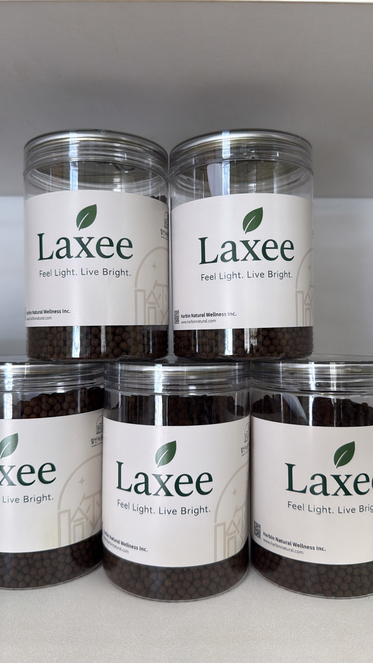 Five clear cylinder jars of Laxee herbal pellets stacked, with the leaf logo and 'Feel Light. Live Bright.' tagline visible
